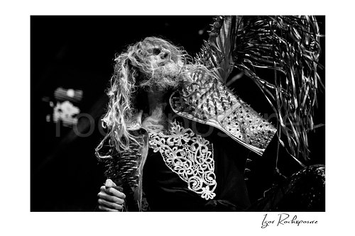 Horizontal black and white image of Rob Zombie performing live on stage with long dreadlocks, a studded fringed jacket, raised arm, and a handheld microphone