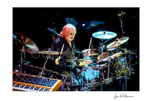 Horizontal color image of Alan White of Yes performing at the drum kit with drumsticks raised during a live performance
