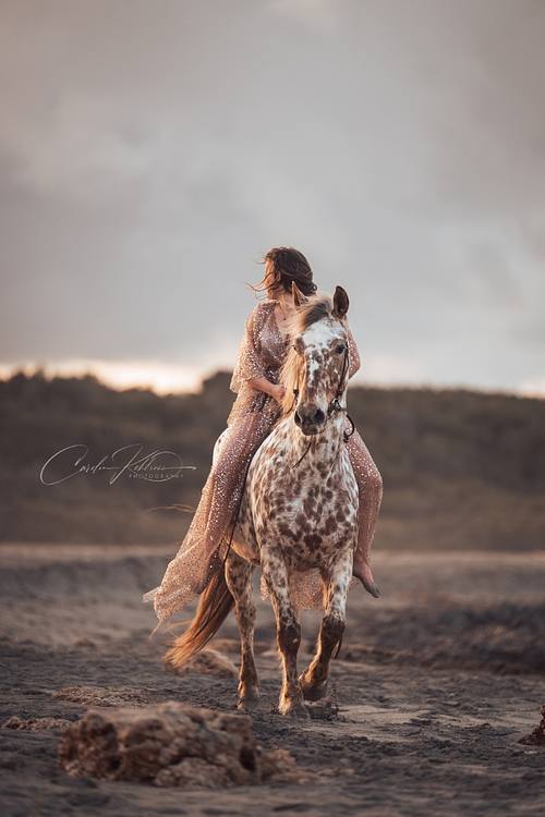 Frau im Paliettenkleid reitet auf Appaloosa am Strand entlang