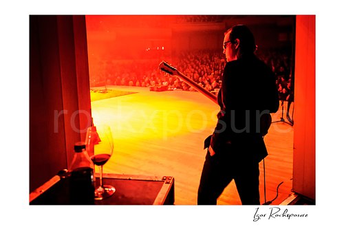 Horizontal color image of Joe Bonamassa standing in the wings before stepping onto the red-lit stage, with a wine glass and bottle of diet coke on a case in the foreground