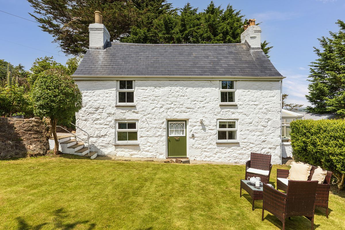 A small white stone cottage with a gray slate roof, green door, three windows, and wicker patio furniture on a grassy lawn, surrounded by trees and shrubs.