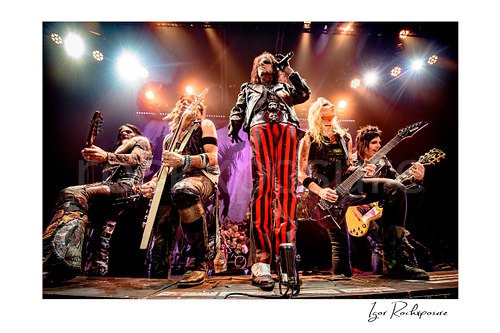 Horizontal color image of Alice Cooper, Chuck Garric, Nita Strauss, Tommy Henriksen and Ryan Roxie of the Alice Cooper Band performing live on stage with striped pants, guitars, and bright concert lighting