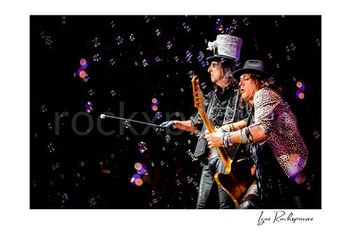 Horizontal color image of Alice Cooper and Ryan Roxie performing live on stage with a silver top hat, guitar, cane, and bubbles floating through the air