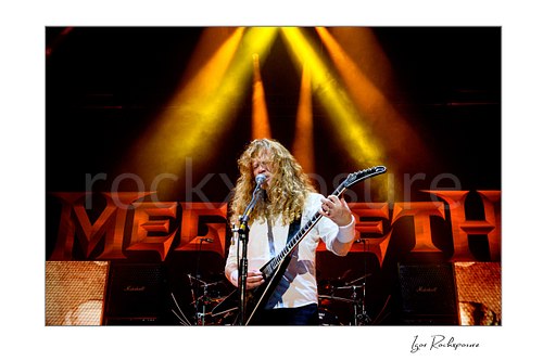 Horizontal color image of Dave Mustaine of Megadeth singing and playing guitar on stage beneath dramatic yellow spotlights