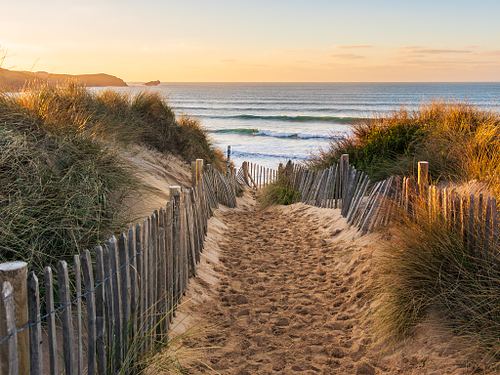 A sandy path through sand dunes lined with wooden fences and tall grass leads to a beach with gentle waves at sunset.