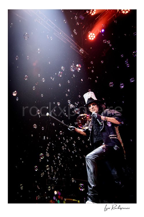 Vertical color image of Alice Cooper performing live on stage in a top hat with a cane, black leather outfit, and bubbles floating through the air