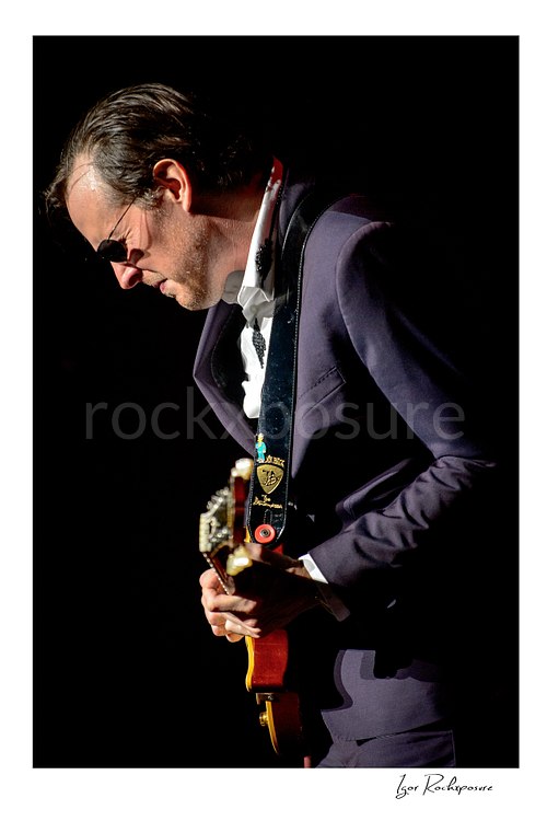 Vertical color image of Joe Bonamassa performing live on stage with his guitar in a dramatic close concert portrait