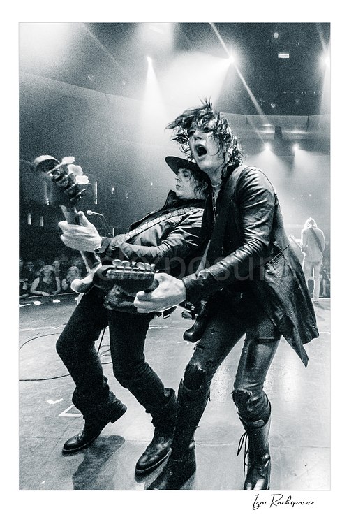 Vertical black and white image of Tom Keifer and Billy Mercer performing live on stage in a dramatic close concert moment, leaning into each other with guitars under bright overhead lights