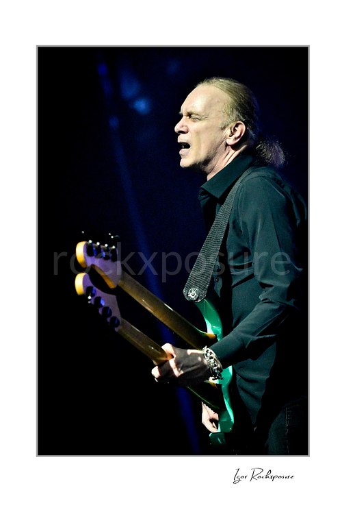 Vertical color image of Billy Sheehan performing live on stage with a double-neck bass guitar, black shirt, and blue concert lighting