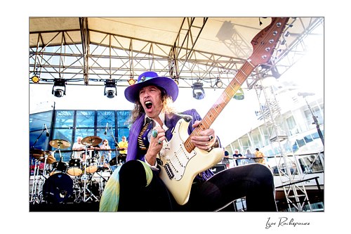 Horizontal color image of Randy Hansen playing a white guitar on an outdoor ship deck stage while wearing a purple hat and jacket