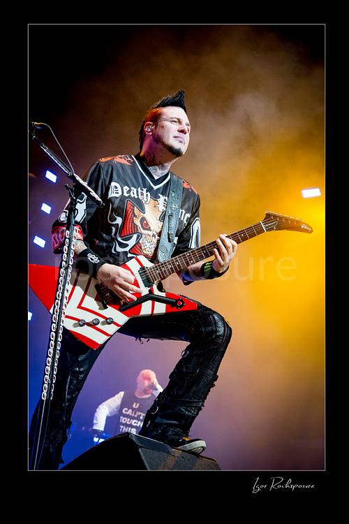 Vertical color image of Zoltan Bathory of Five Finger Death Punch playing guitar onstage under warm yellow concert haze