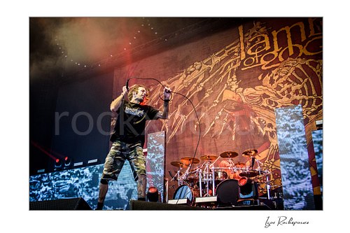 Horizontal color image of Randy Blythe of Lamb of God performing on stage with a handheld microphone, looping cable, and the drum kit behind him