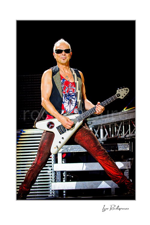 Vertical color image of Rudolf Schenker of Scorpions performing live with white guitar, sunglasses, and red stage pants