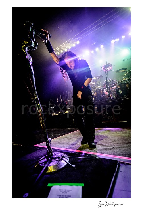 Vertical color image of Korn singer Jonathan Davis standing on stage beside his distinctive stage microphone stand with the drum kit visible behind him