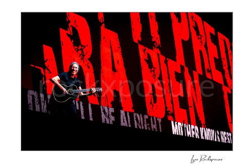 Roger Waters plays a black acoustic guitar at the left of a wide stage view dominated by oversized red lettering and smaller white text.