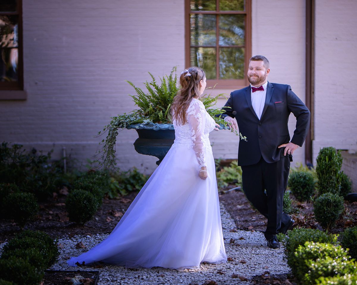 groom posing in the formal garden at ross mansion with the bride slowly walking towards him