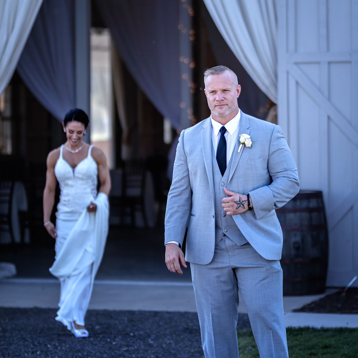 first look at kylan barn, the groom is standing, holding his hand to his jacket, waiting for the bride to approach from the barn