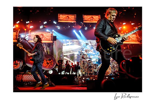 Horizontal color image of Rush performing live on stage with Geddy Lee, Neil Peart, and Alex Lifeson under vivid red and blue concert lighting