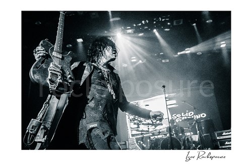 Horizontal black and white image of Tom Keifer on stage holding his guitar upright in a dramatic live performance moment, with bright concert beams and the drum kit visible behind him