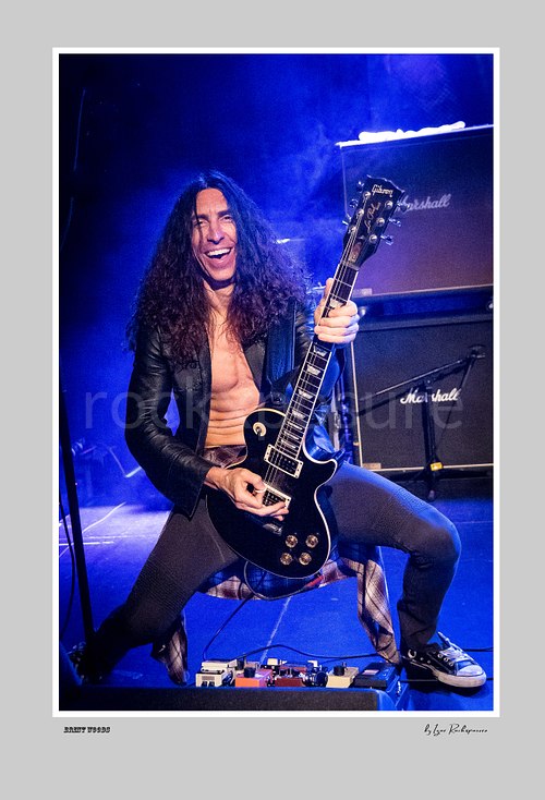 Vertical color image of Brent Woods playing guitar on stage with a wide smile, open jacket, and blue stage light