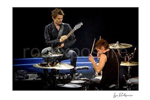 Horizontal color image of Matt Bellamy and Dominic Howard of Muse performing live on stage with guitar and drums in a dramatic concert moment