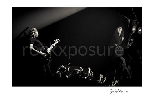 Black and white stage photograph of Roger Waters playing bass beneath a diagonal beam, with raised hands below and a projected character at right.