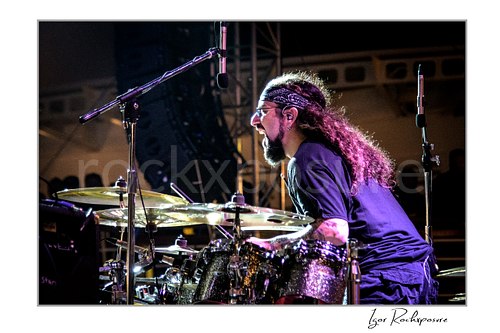 Horizontal color image of Mike Portnoy at the drum kit in side profile under purple stage lighting with glowing hair