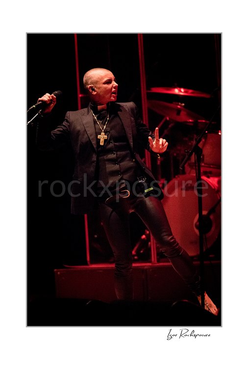 Sinéad O’Connor turns sideways with a microphone lifted near her shoulder and one finger raised, wearing a black jacket before a red-lit drum kit.