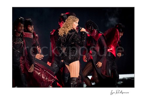 Horizontal color image of Taylor Swift singing on stage in profile with dancers and red concert lighting