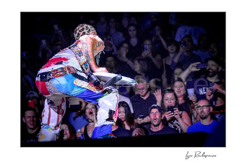 Flea crouches facing the front row with his bass across his thighs, patchwork trousers and tattooed back visible as fans watch and raise phones.