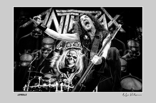 Horizontal black and white image of Frank Bello of Anthrax performing live on stage, raising one arm while playing bass guitar in front of the band’s skull-themed stage backdrop