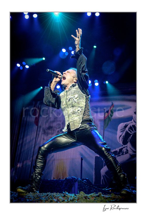 Vertical color image of Bruce Dickinson of Iron Maiden performing live on stage, singing into a microphone in a dramatic wide stance with one arm raised under blue concert lights.