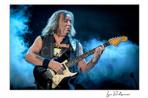 Horizontal color image of Dave Murray of Iron Maiden performing live on stage, playing electric guitar against a blue smoky concert background.