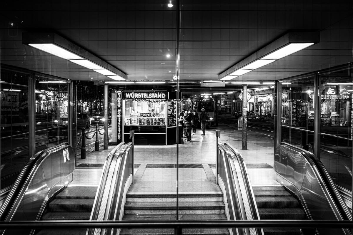 Schwarz-Weiß-Fotografie eines Wiener Würstelstands bei Nacht hinter Rolltreppenabgängen mit symmetrischer Architektur und urbaner Beleuchtung