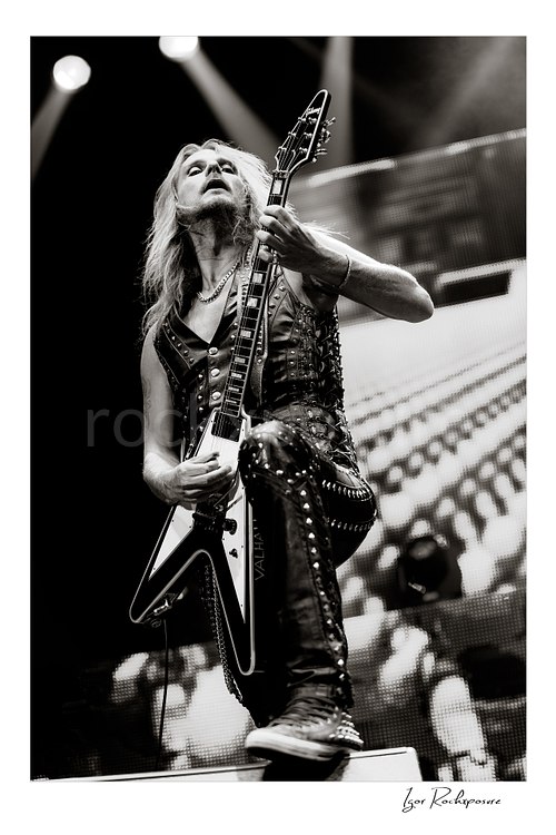 Vertical black and white image of Richie Faulkner of Judas Priest performing live on stage, playing flying V guitar in a dramatic low-angle shot under bright concert lighting