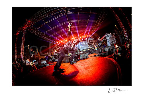 Horizontal color image of Joey Tempest of Europe performing live on stage in a crouched pose with a raised microphone, wide fisheye perspective, and vivid red concert lighting