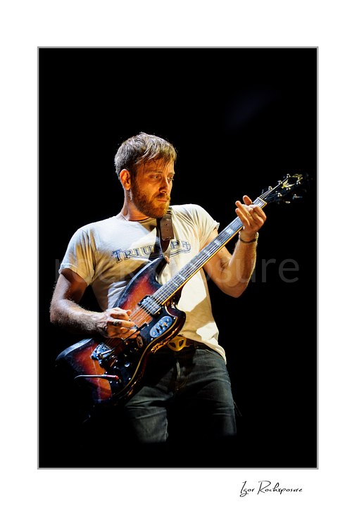 Vertical color image of Dan Auerbach of The Black Keys playing guitar under warm stage lighting in a dark concert scene