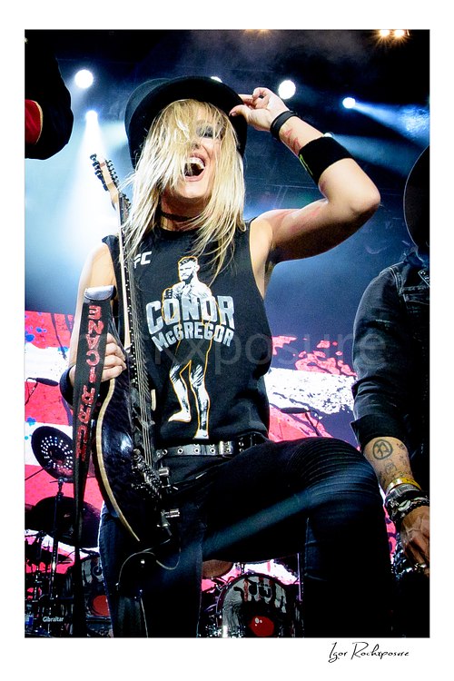 Vertical color image of Nita Strauss performing live on stage with an electric guitar, smiling while wearing a black hat and a Conor McGregor shirt