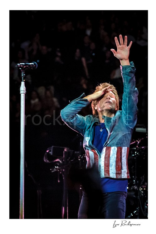 Vertical color image of Jon Bon Jovi on stage with one arm raised and the microphone stand beside him during a live performance