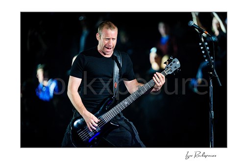 Horizontal color image of Mike Kroeger of Nickelback playing bass onstage beside a microphone stand during a live performance