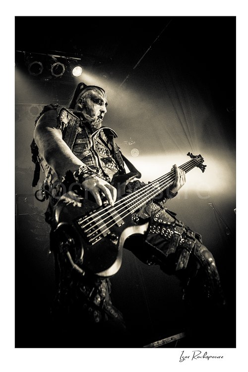Vertical black and white image of Orion of Behemoth playing bass in a dramatic low-angle live performance shot under bright stage backlight