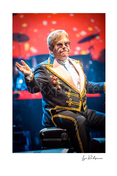 Vertical color image of Elton John seated at the piano with arms raised before a glowing red stage backdrop