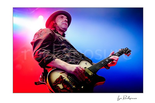 Horizontal color image of Phil Campbell of Motörhead performing live on stage with an electric guitar, brimmed hat, and vivid red and blue concert lighting