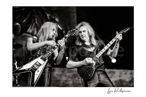 Horizontal black and white image of Richie Faulkner and Glenn Tipton of Judas Priest performing live on stage, with Faulkner at left playing flying V guitar and Tipton at right playing electric guitar