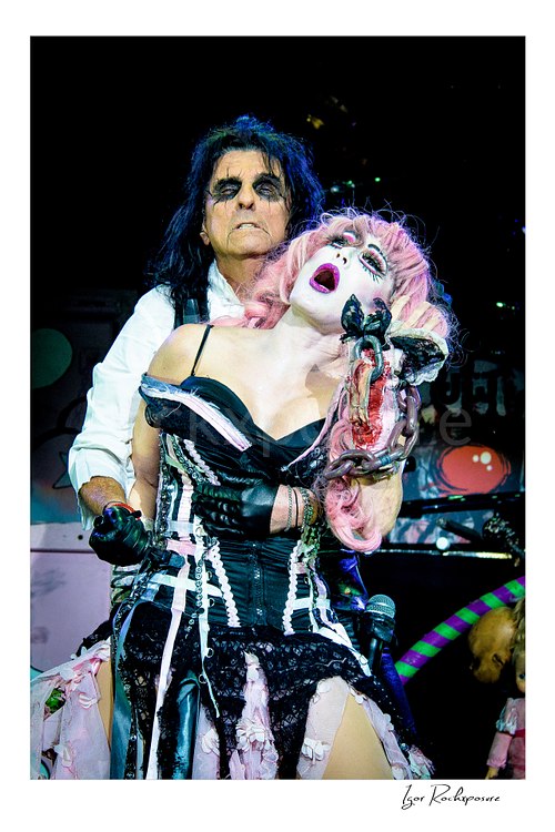 Vertical color image of Alice Cooper and Sheryl Cooper performing live on stage in theatrical makeup and costume with pink hair, black corset, and a dramatic horror prop
