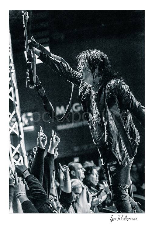 Vertical black and white image of Tom Keifer performing live at the edge of the stage, raising his black guitar overhead toward the crowd in a fierce concert moment