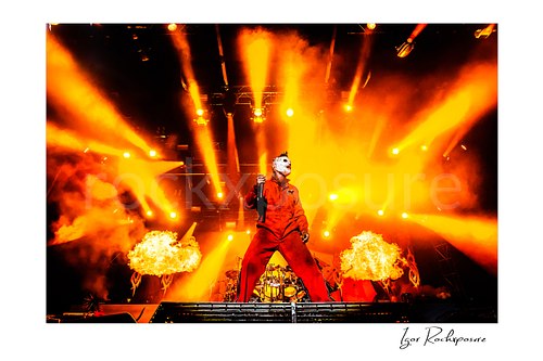 Horizontal color image of Corey Taylor of Slipknot standing center stage in a red jumpsuit and mask with a handheld microphone, fire bursts, and blazing orange concert lights