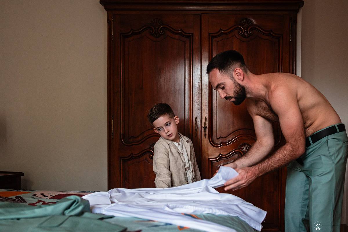 Dans une chambre avec une armoire en bois classique, un homme torse nu ajuste des v&ecirc;tements sur un lit tandis qu'un jeune gar&ccedil;on en costume regarde attentivement, symbolisant la pr&eacute;paration intime avant la c&eacute;r&eacute;monie la&iuml;que sous la tente du mariage &agrave; N&icirc;mes