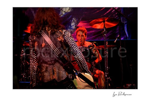 Horizontal color image of Jan LeGrow and Sean Sellers of Beasto Blanco performing live on stage, with LeGrow seen from behind in the foreground and Sellers at the drum kit under vivid red stage light