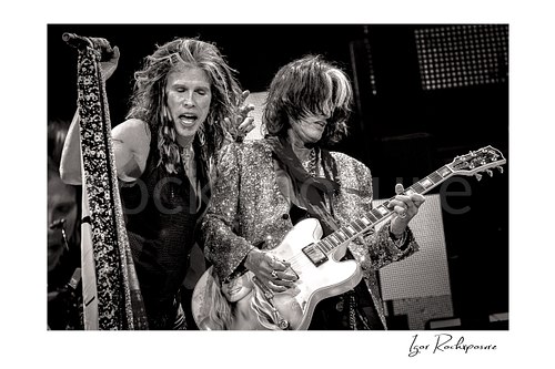 Horizontal black and white image of Steven Tyler and Joe Perry of Aerosmith performing live on stage with a scarf-wrapped microphone, glitter jacket, and electric guitar
